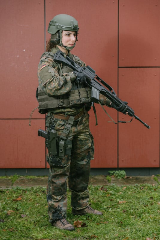 A soldier in camouflage uniform and helmet stands on grass in front of a red wall, holding a rifle with both hands and looking forward. The soldier is wearing tactical gear and boots.