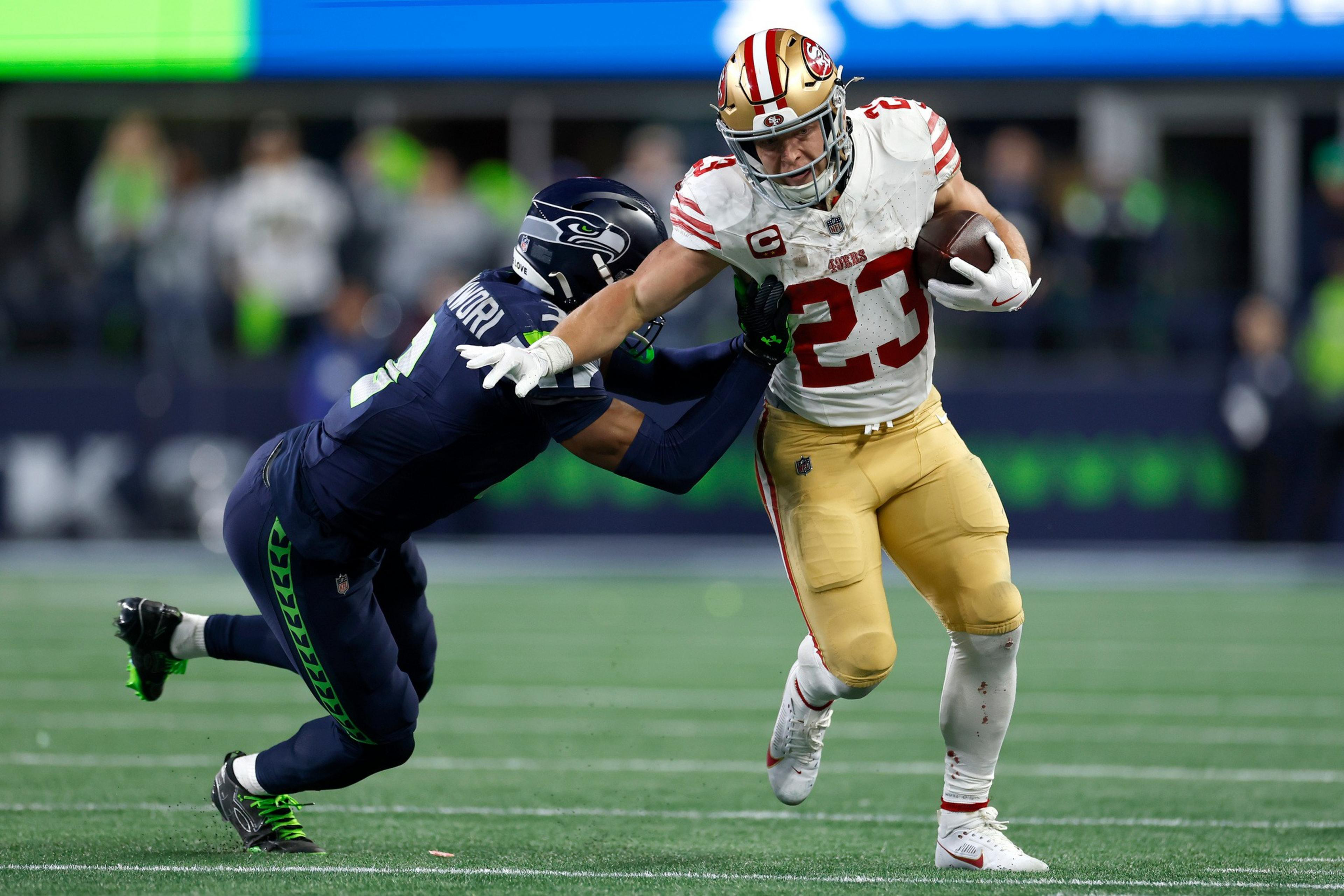 A football player in a white and gold uniform carries the ball while pushing away a defender in a dark blue uniform attempting to tackle him.