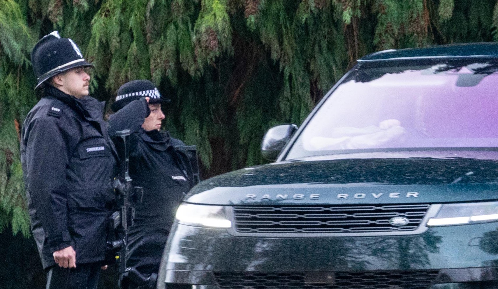King Charles III leaving Sandringham Estate in a dark SUV with two police officers standing beside the car.