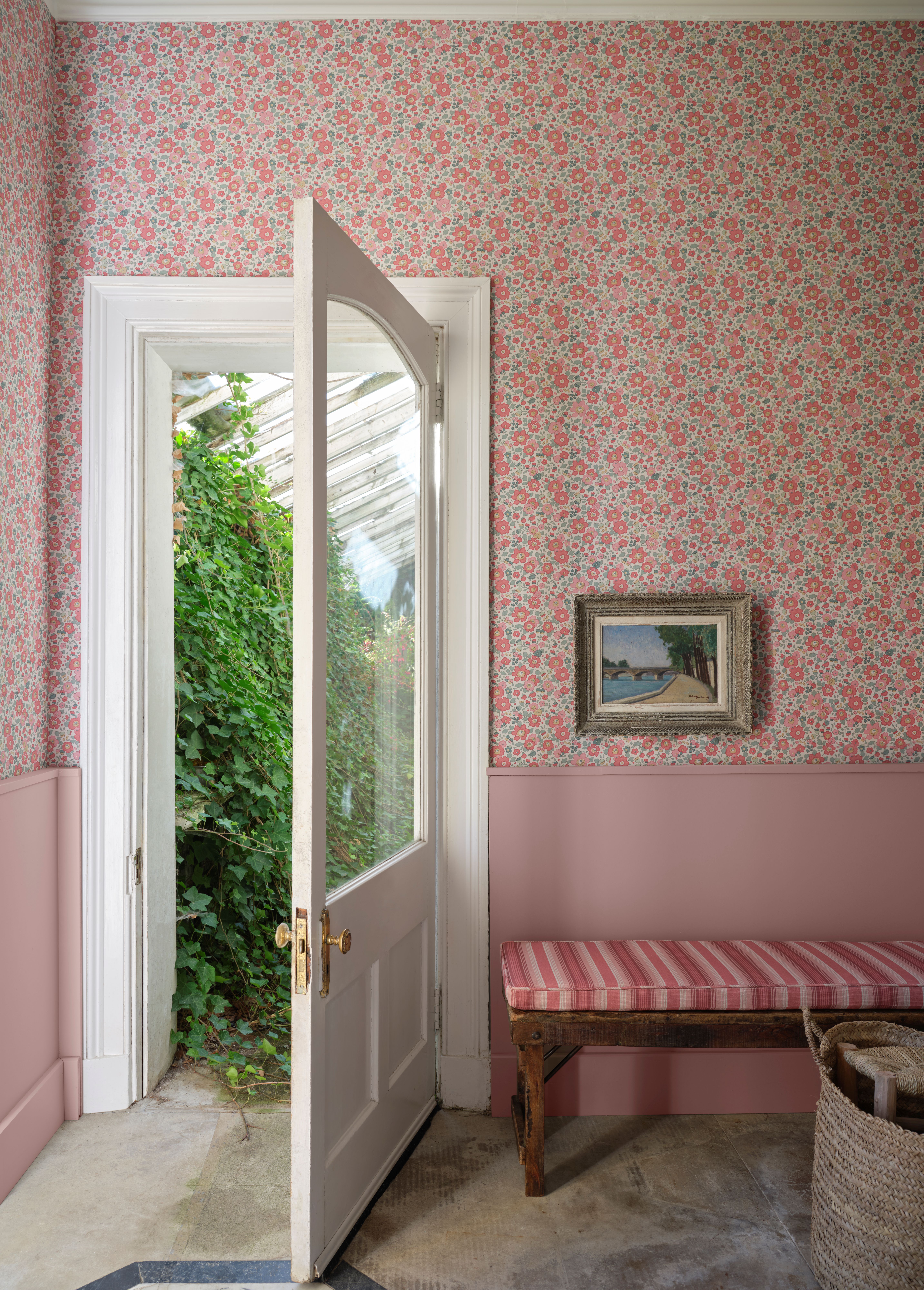 Room with pink floral wallpaper, a painted pink dado rail, a striped bench, and an open door revealing an ivy-covered greenhouse.
