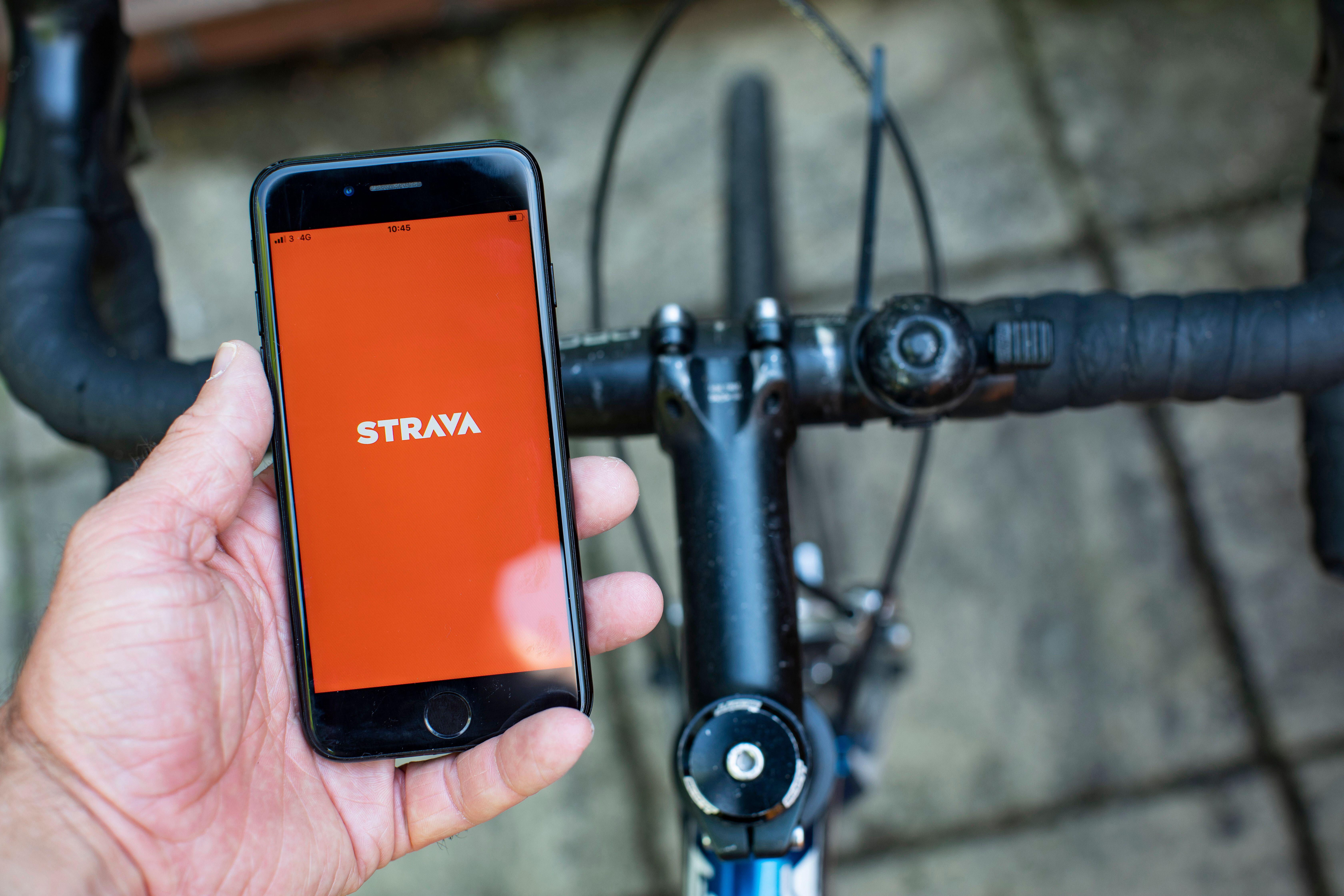 A hand holding an iPhone displaying the Strava app next to a bicycle's handlebars.