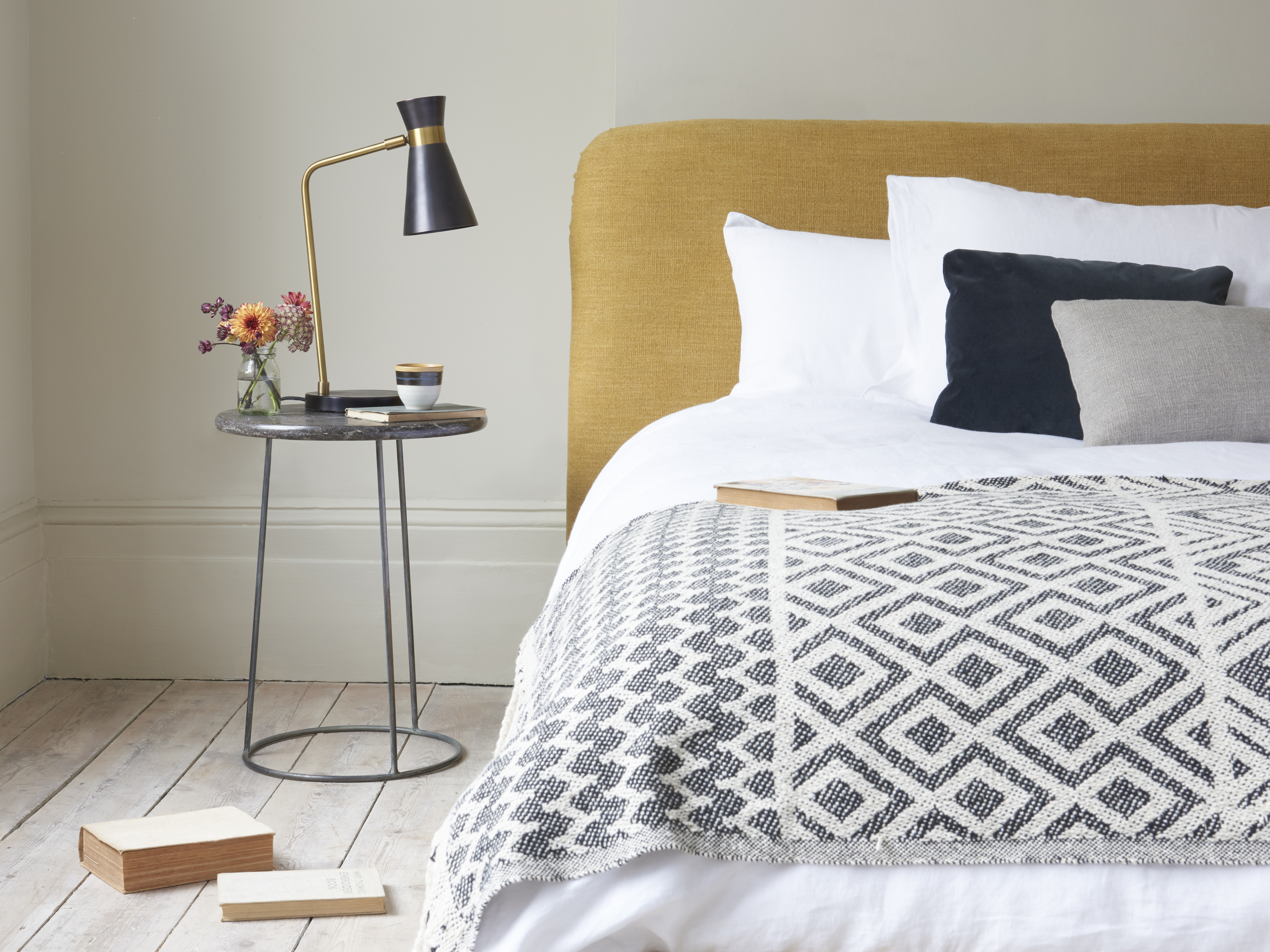 A neatly made bed with a mustard-yellow headboard and a patterned throw blanket, next to a marble nightstand with a lamp and flowers.