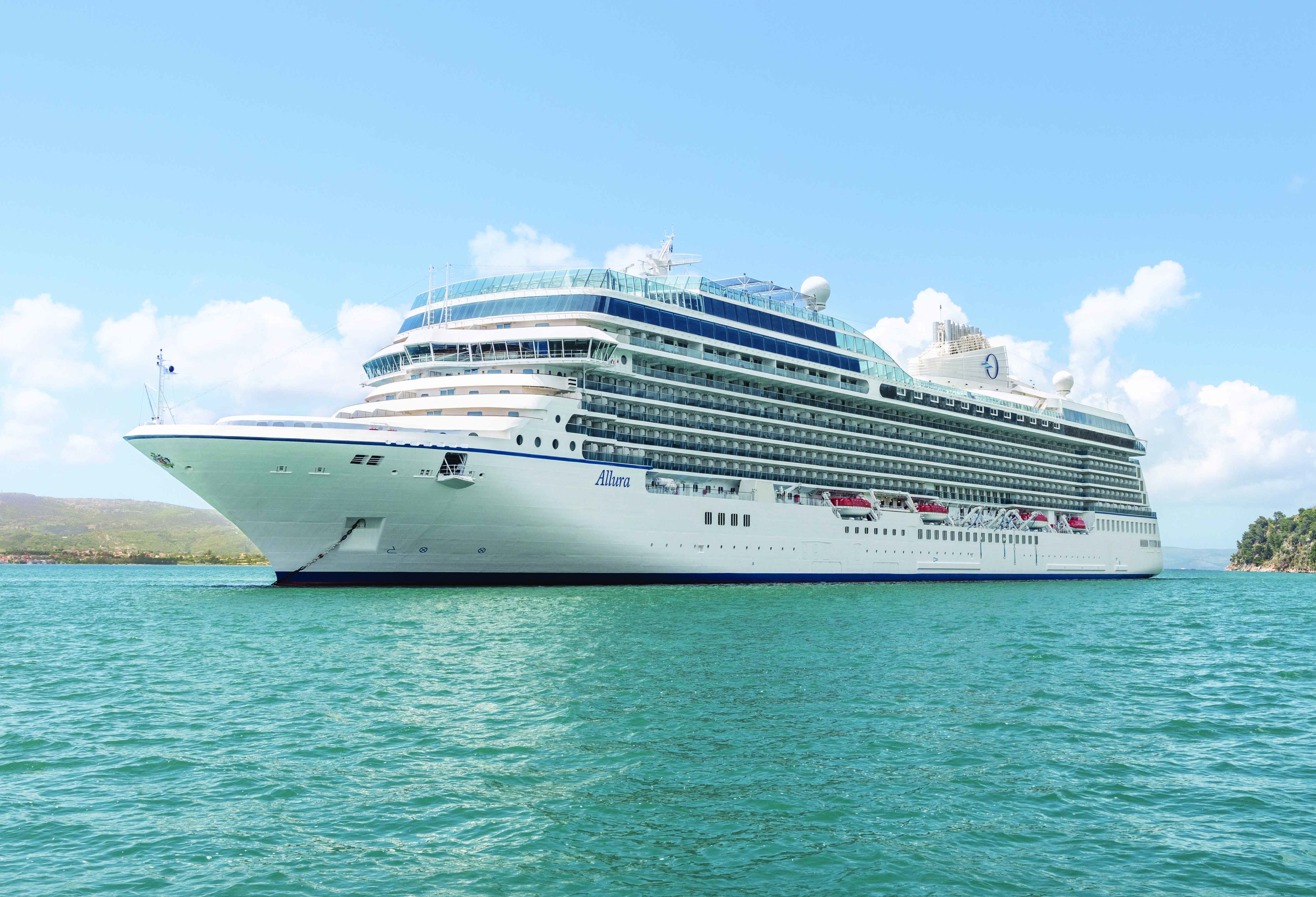 Oceania Allura cruise ship sailing on turquoise water with a mountainous landscape in the background.