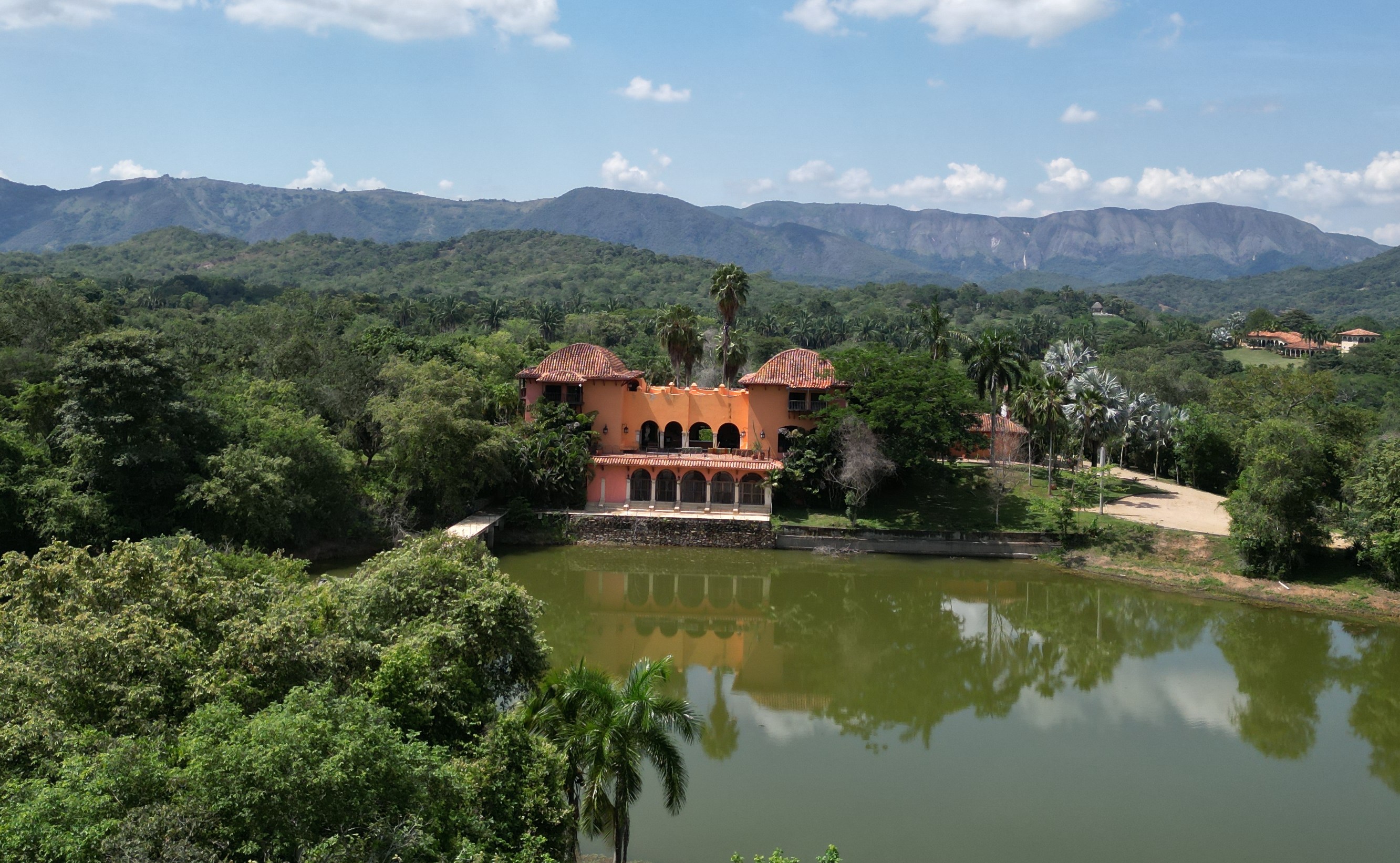 Roper's Villa, a large orange building with red tiled roofs, sits beside a green lake, surrounded by dense trees and distant mountains under a blue sky with white clouds.