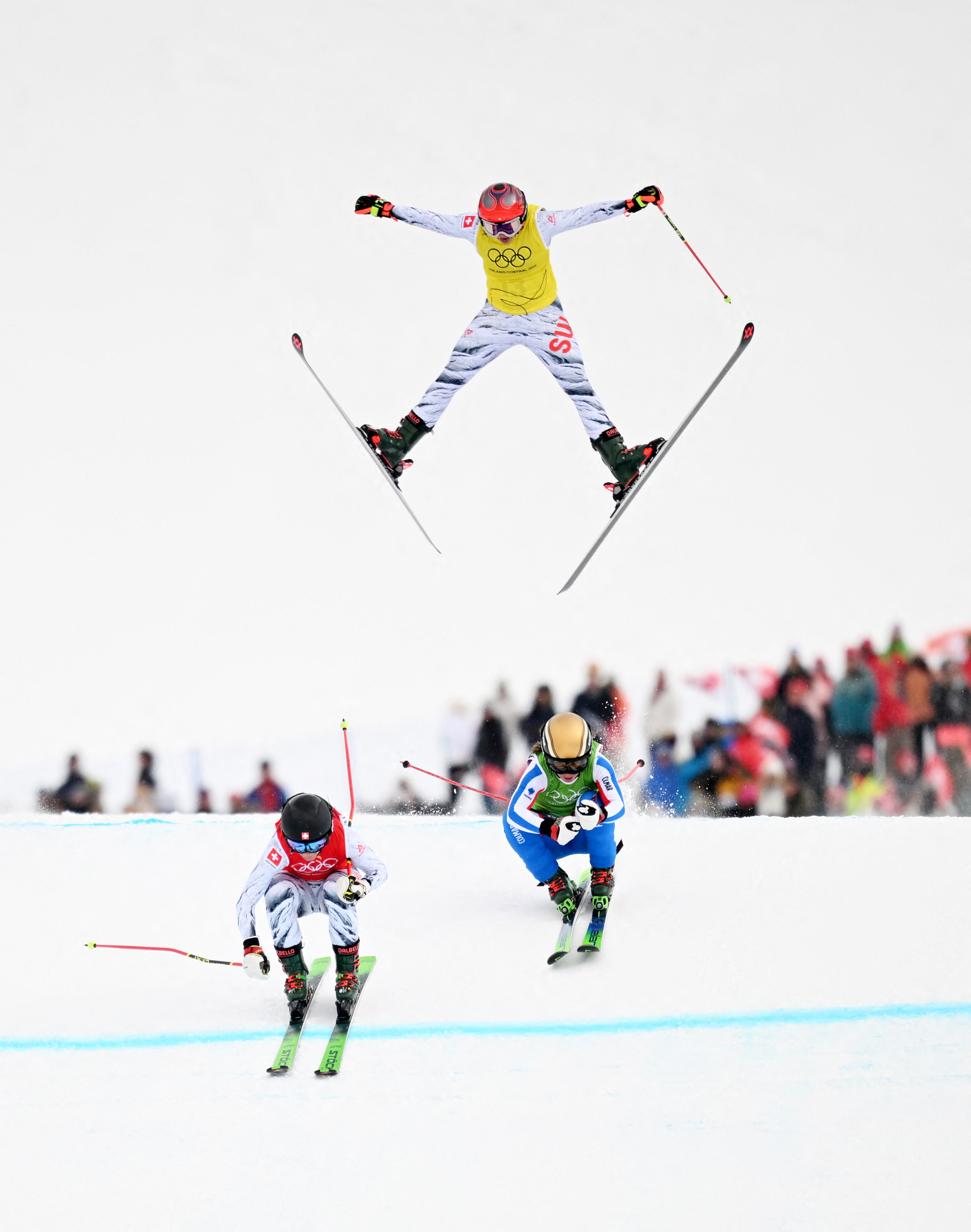 Three female freestyle skiers racing, one airborne and two close behind.