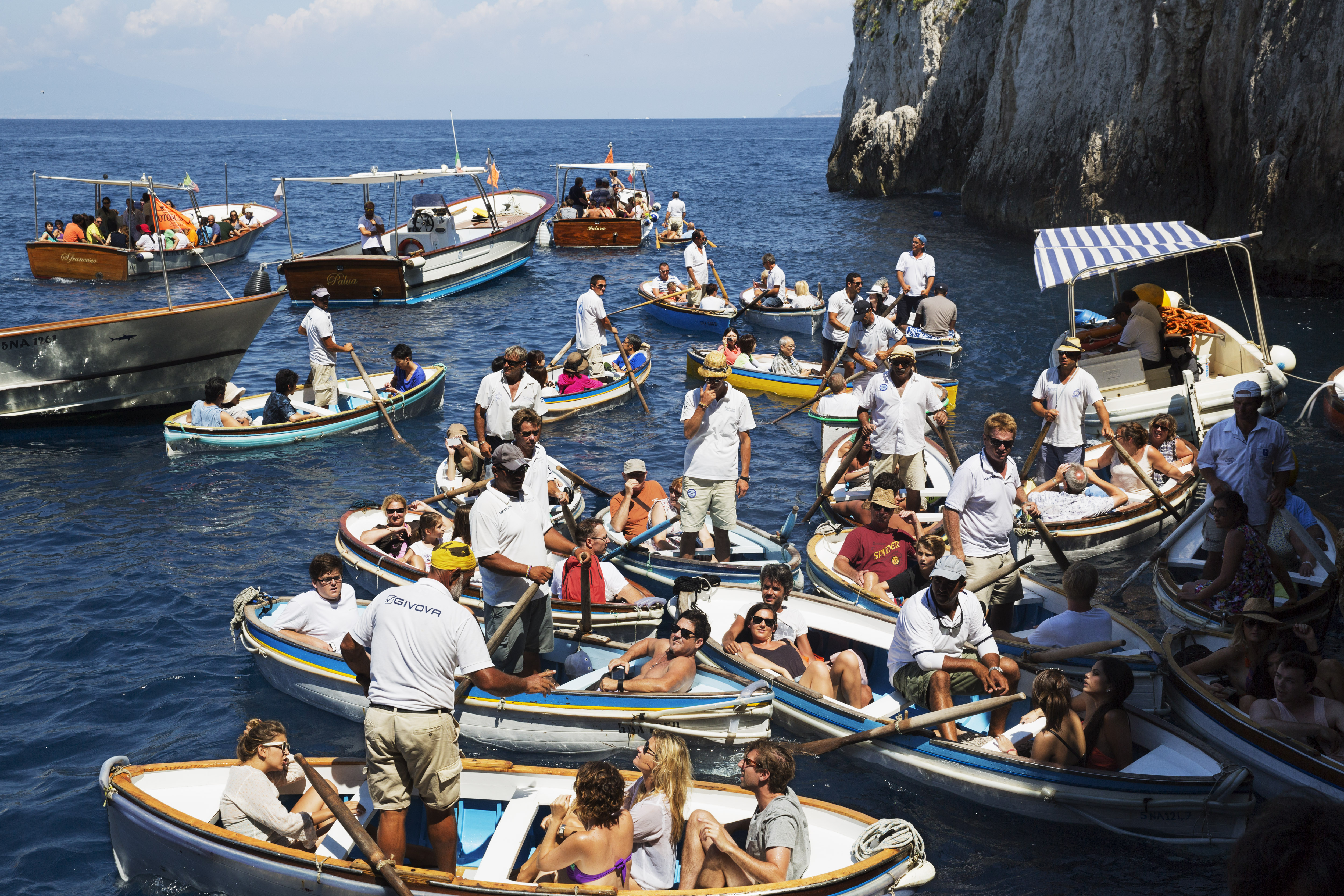 People in small boats, including rowboats and motorboats, wait to enter the Blue Grotto in Capri, Italy.