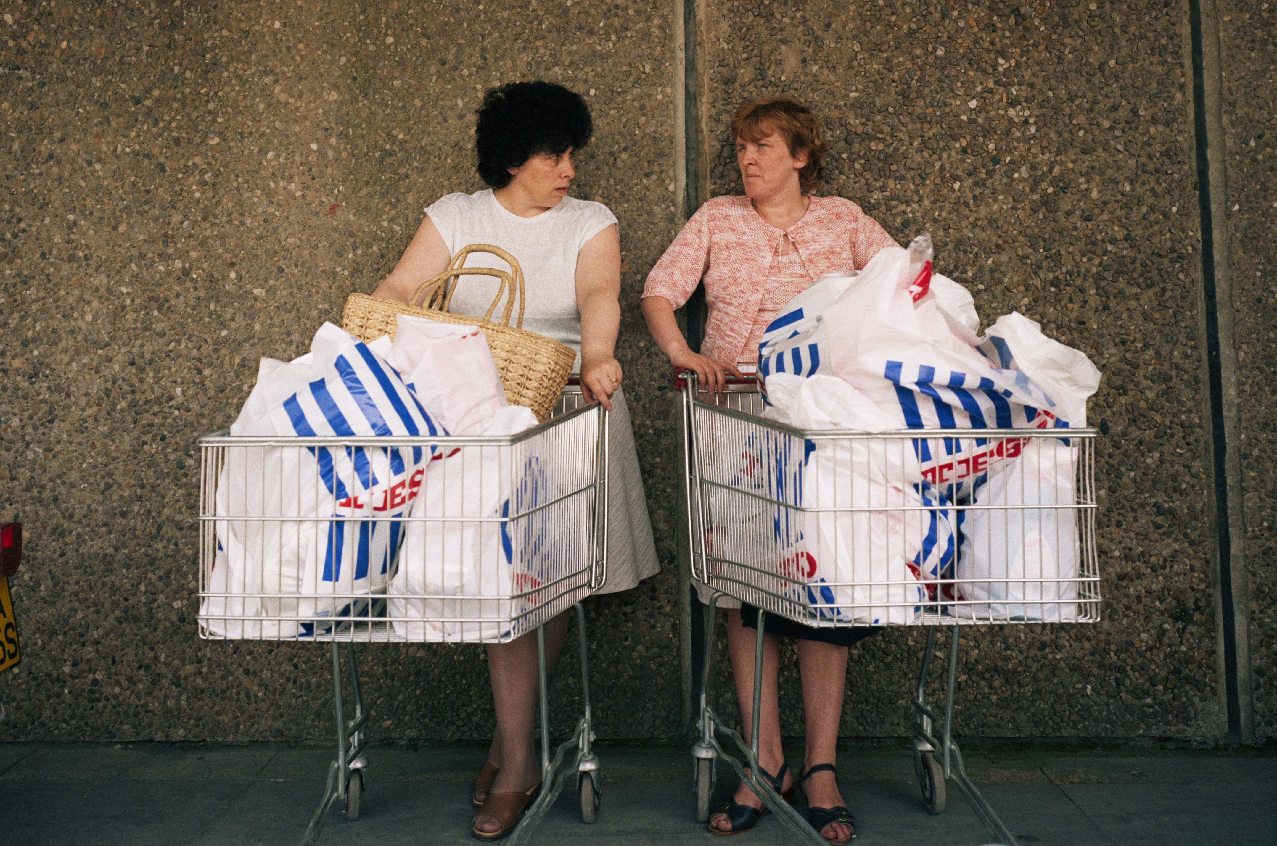Two women stand with shopping carts full of groceries in front of a stone wall.