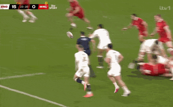 Rugby players in red and white jerseys scramble for the ball on a green field.