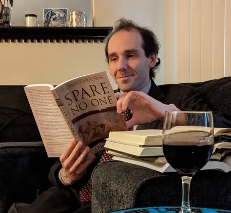 Bret Devereaux, an ancient history PhD, sits on a couch reading "Spare No One."