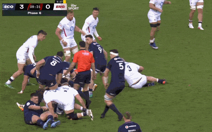 Rugby players in white and dark blue jerseys gathered around the ball on a green field.