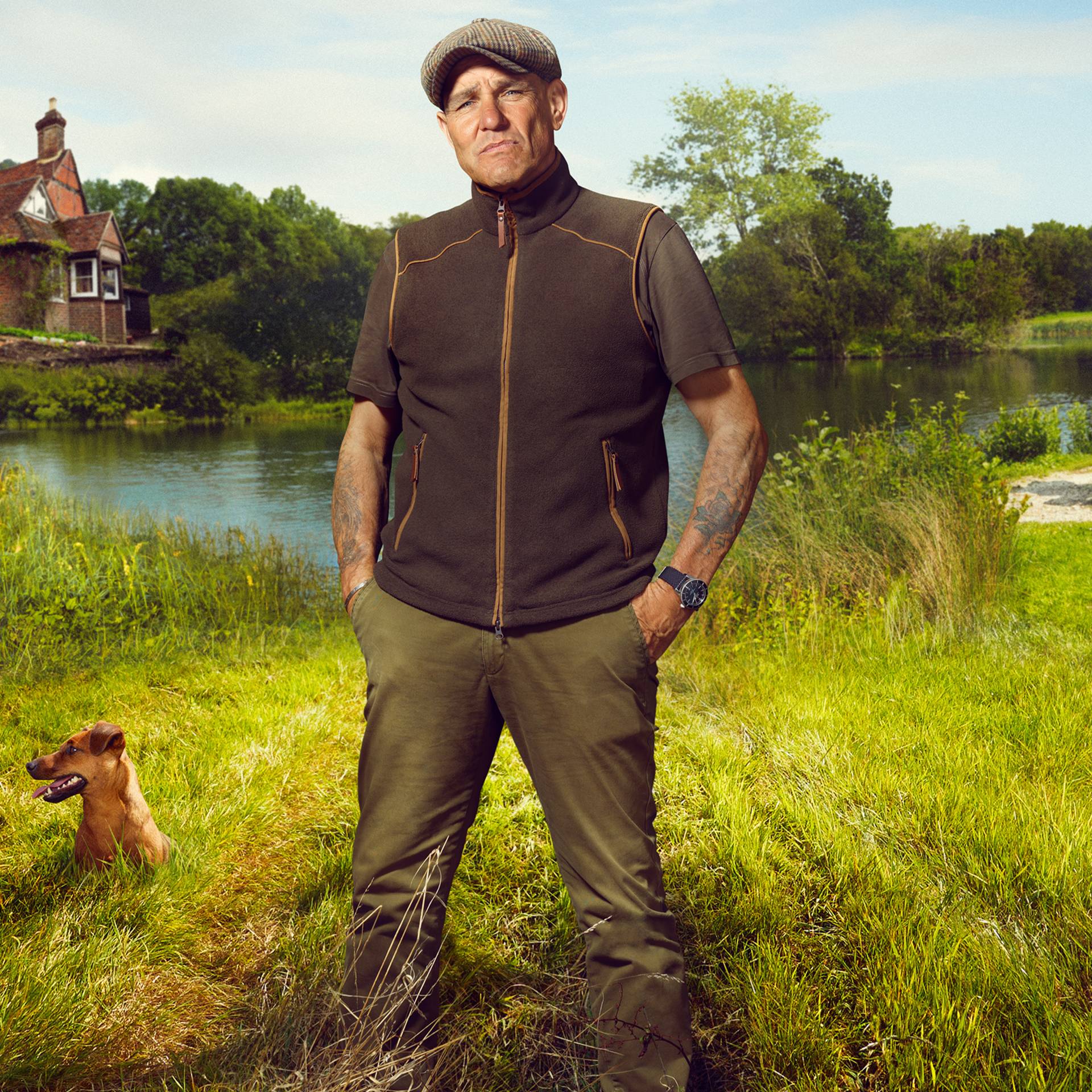 Vinnie Jones standing in a grassy field with a lake and house in the background.