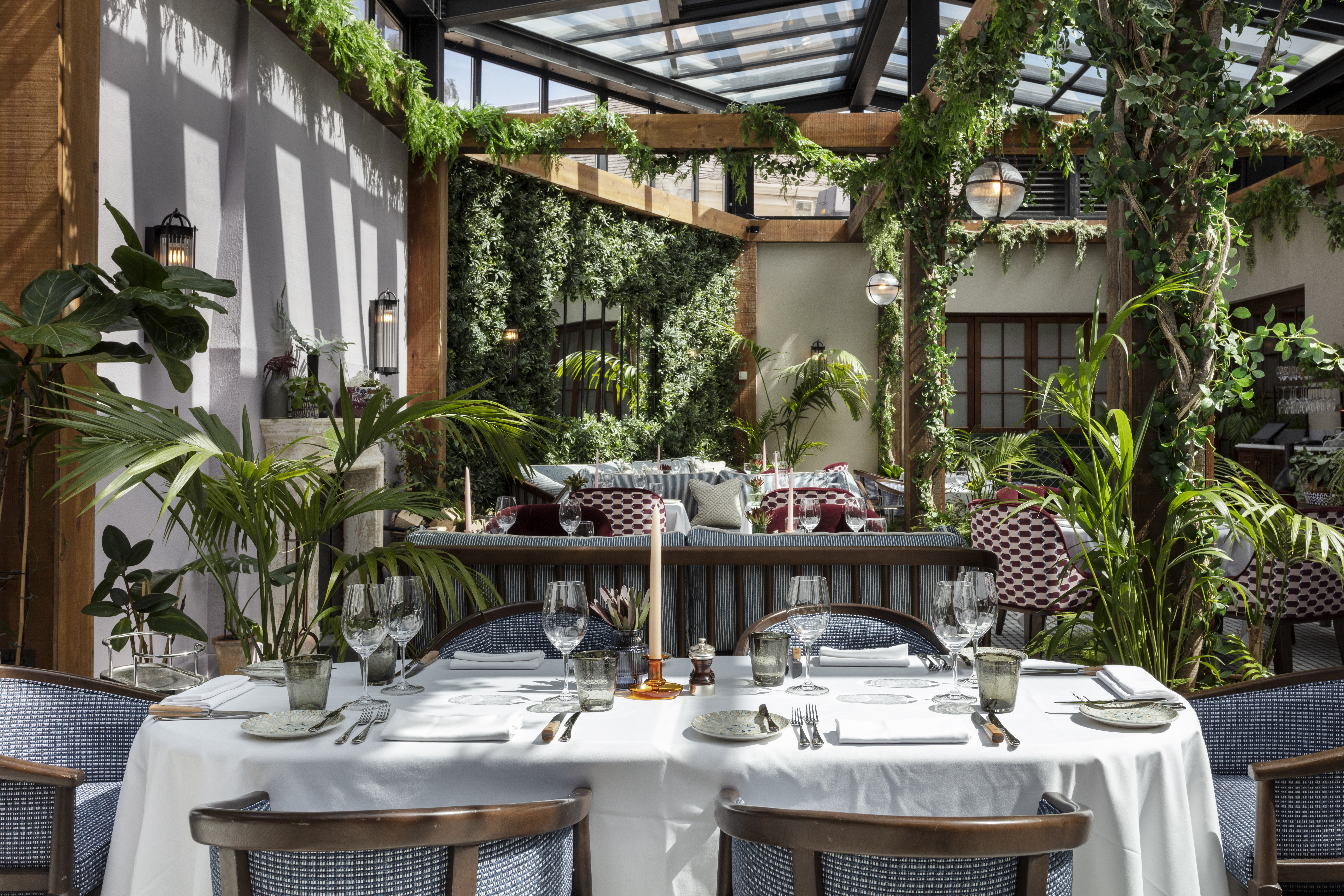 An interior view of a restaurant with tables set and abundant greenery, including a glass ceiling.