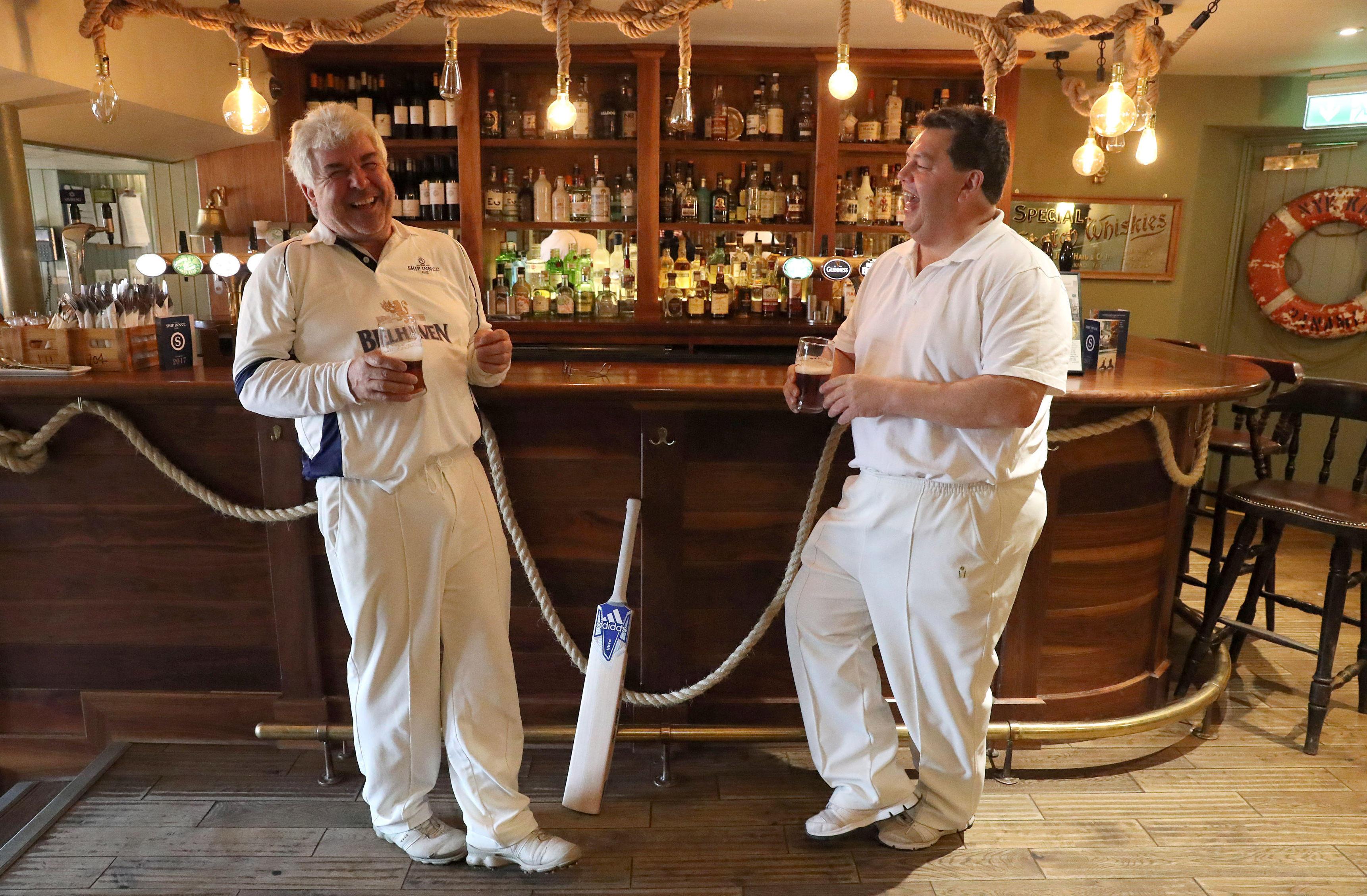 Two men in white cricket uniforms stand at a bar, laughing and holding glasses of beer, with a cricket bat leaning between them.