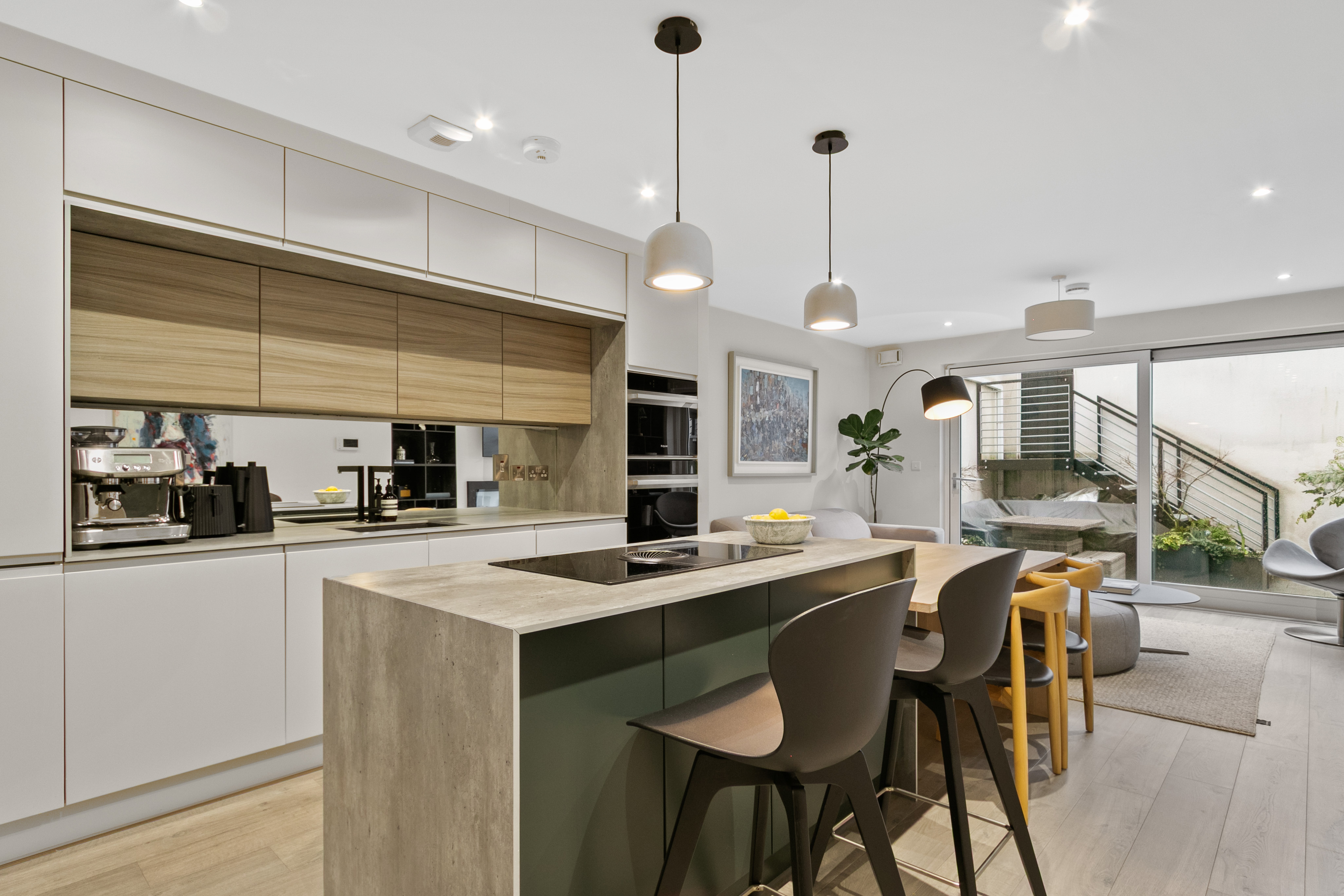 Modern kitchen with light grey and wood grain cabinets, a concrete-look island with a hob, black bar stools, and a dining area.