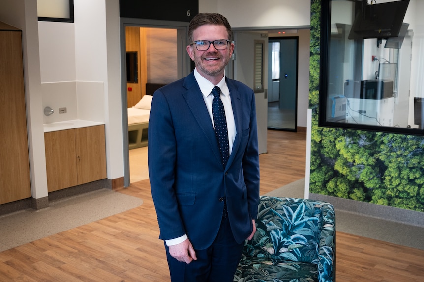A man wears a navy suit, he stands smiling and has eye gasses on with short brown hair. Around him is a new clinical setting.