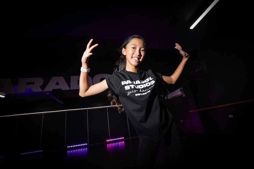 A girl with a dark tshirt in a dark room smiling at the camera with hands raised