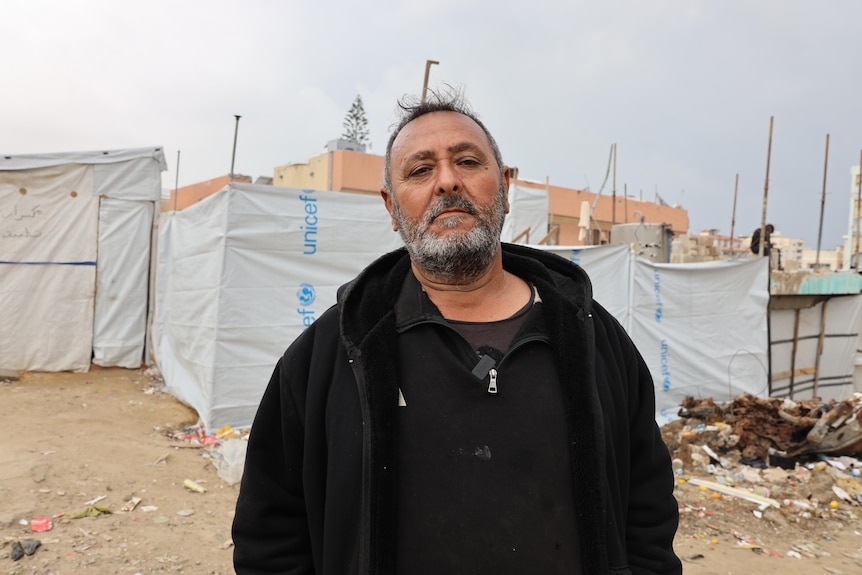 A man wearing black clothing with a grey beard standing in front of a dishevelled UNICEF tent