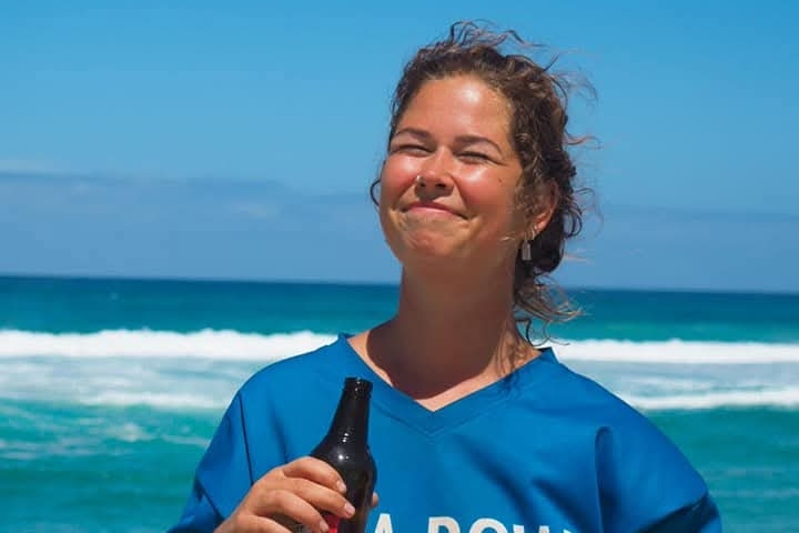 A woman in a blue shirt smiles into the sun, holding a bottle and ocean waves crashing behind her.
