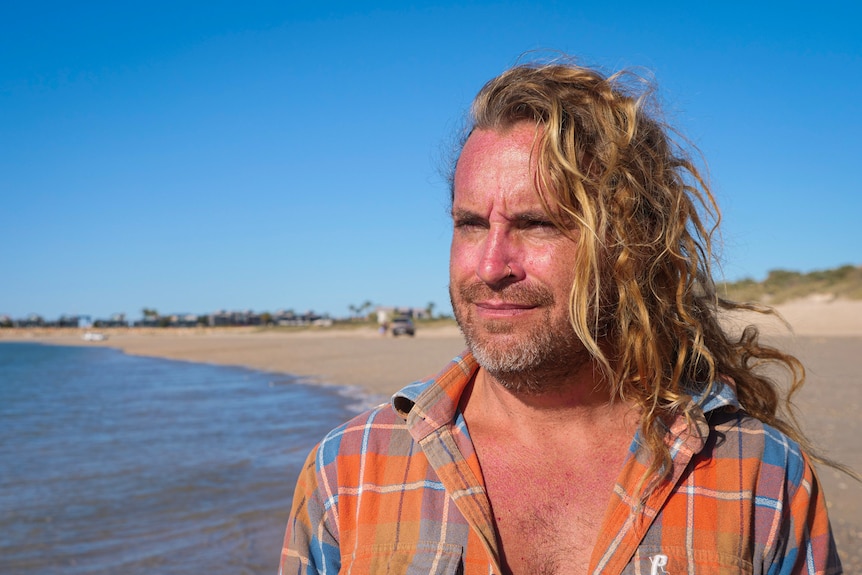 A man with long blonde hair smiles on a beach.