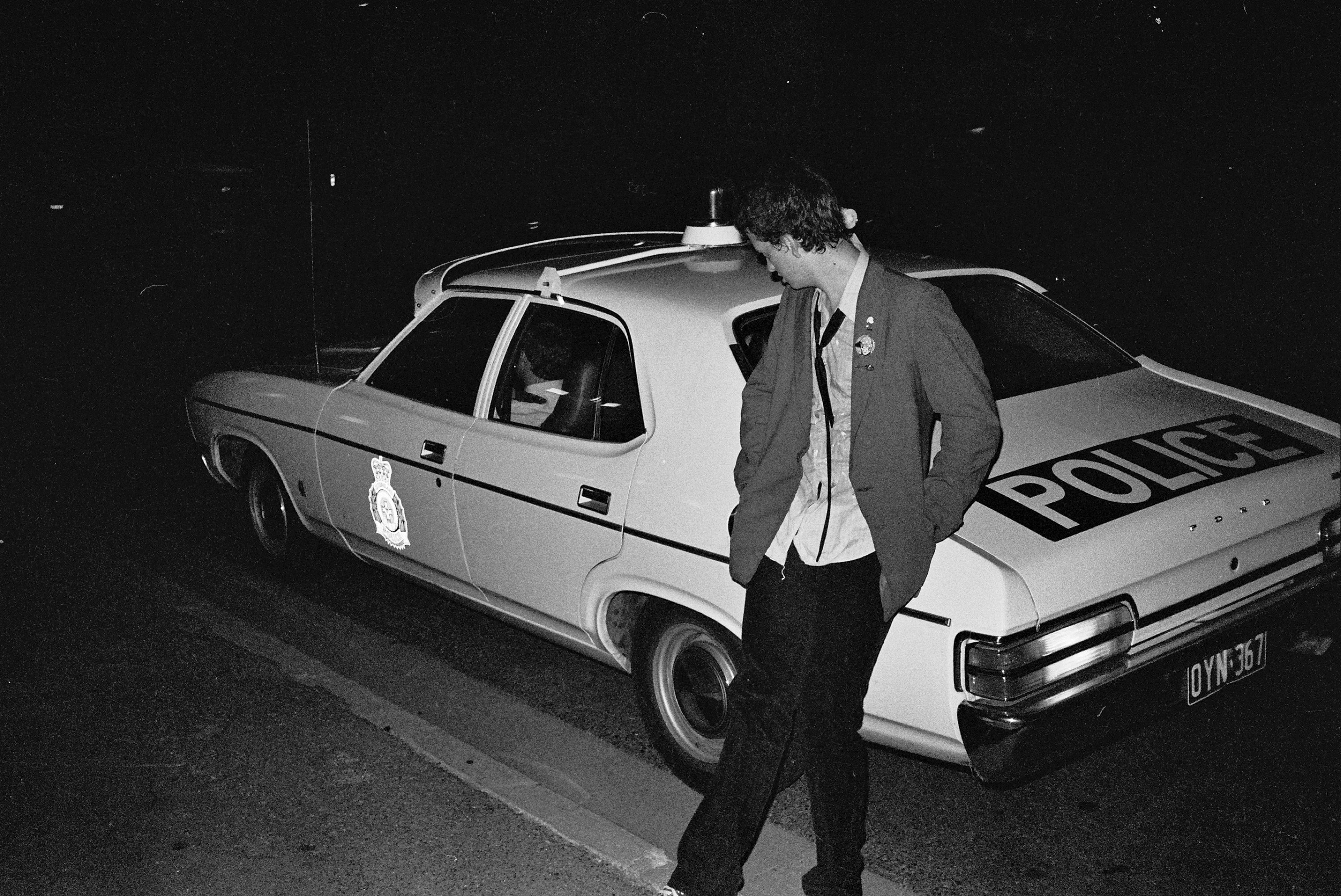 A black and white photo of a man dressed in punk clothing leaning against a police car