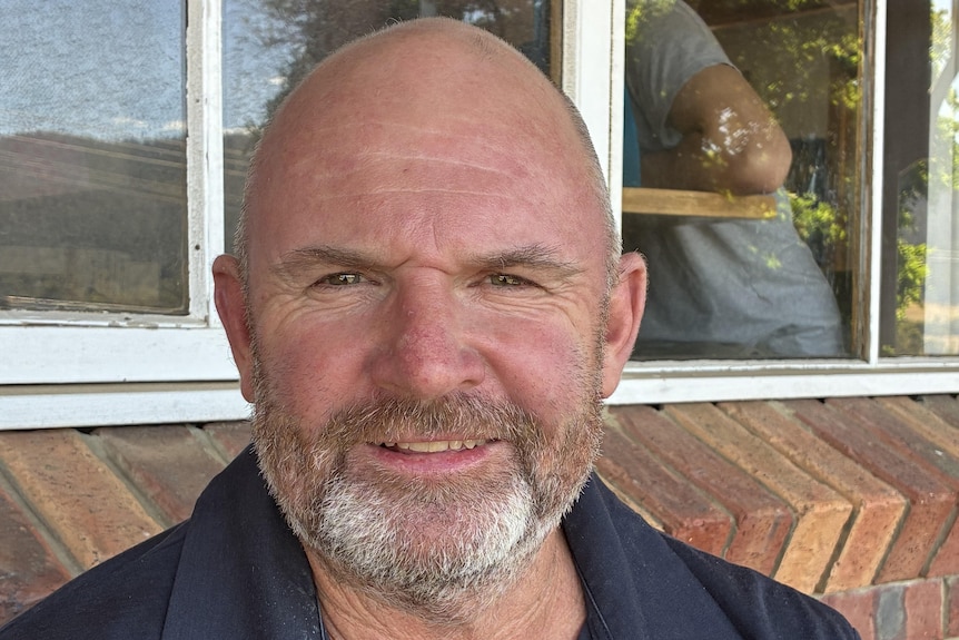 A man in a collared t-shirt sits in front of a brick wall and window.