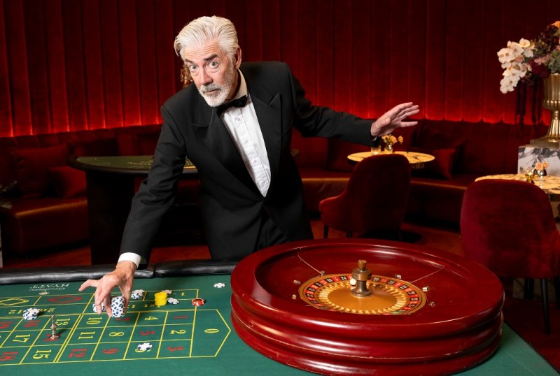 A well-dressed casino dealer in a tuxedo gestures at a roulette table, showcasing vibrant chips and a spinning wheel, set against a luxurious red velvet backdrop.