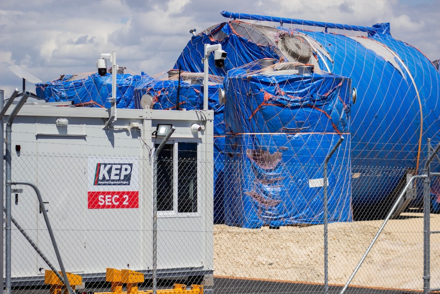 Big mechanical parts and steel wrapped in tarpaulin behind a wire fence next to a small security building