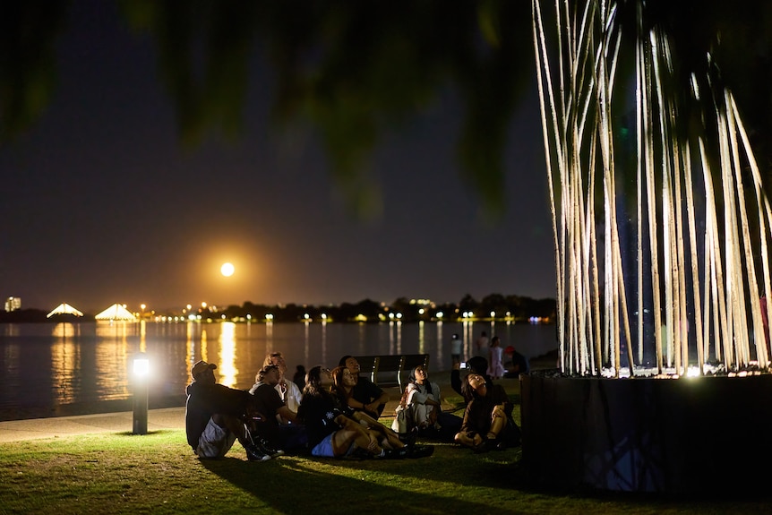 Light sculpture by the river with people sitting on grass looking up 