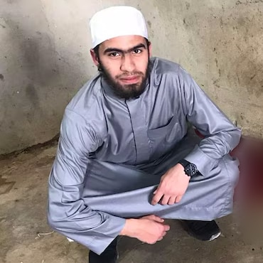 A man wearing a traditional Muslim cap and long shirt crouches on a concrete floor. He is looking at the camera.