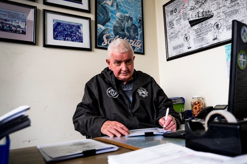 A man with white hair in a black spray jacket and striped shirt sits behind a computer screen at a desk holding a white pen.