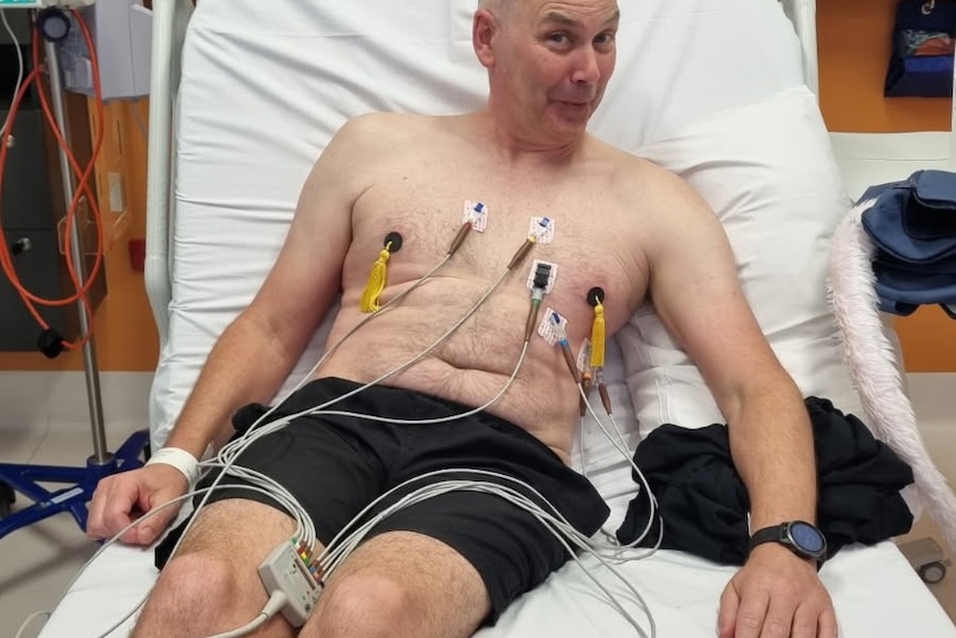 A bald man lying shirtless in a hospital bed, with nipple tassels on.