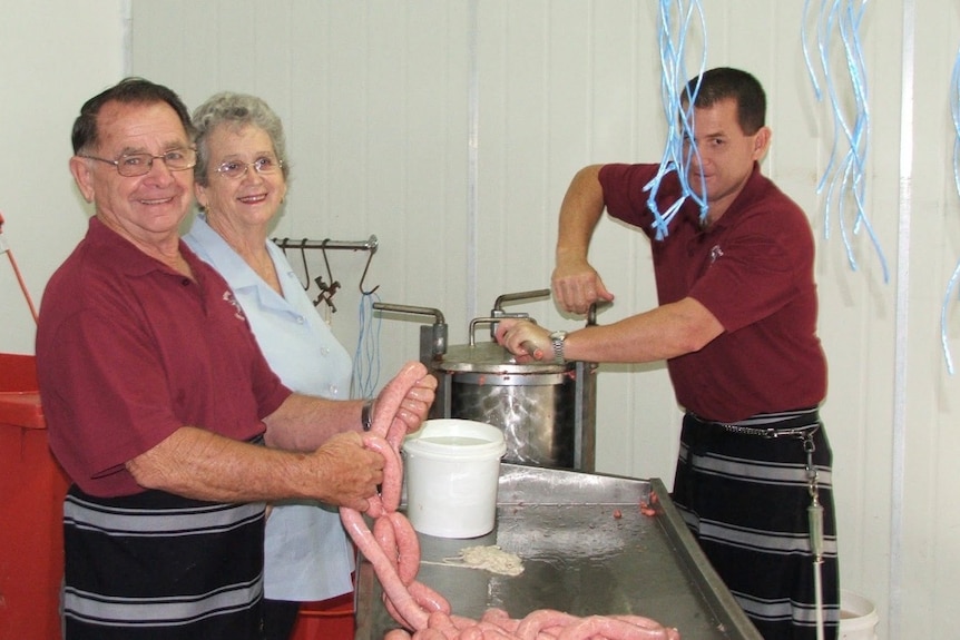An elderly man and woman stand side by side smiling while tying sausages. Across from them their son grinds meat.
