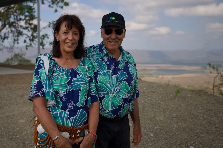A smiling couple stand outdoors on a hilltop with farmland stretching into the distance behind them.