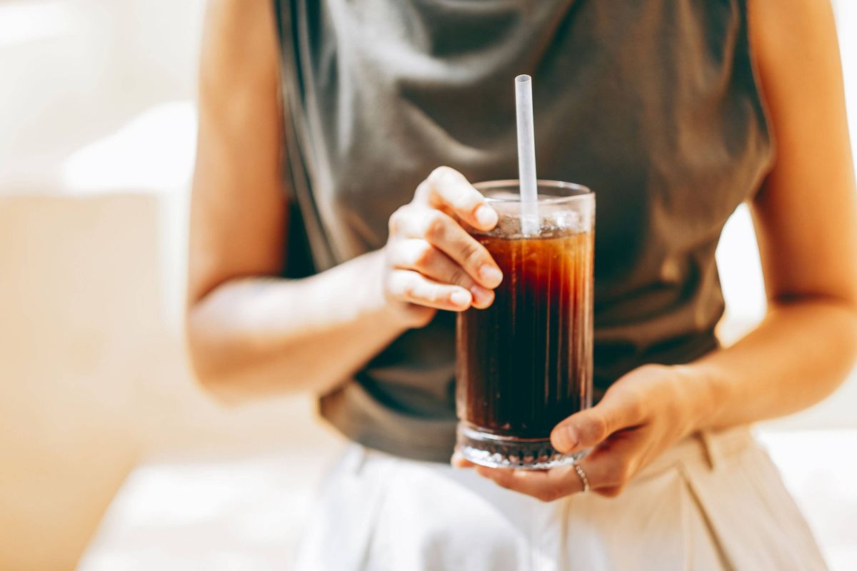 A woman in a sleeveless top and light trousers holds a glass of iced dark soda with a straw. Only her torso and hands are visible. Soft, bright lighti