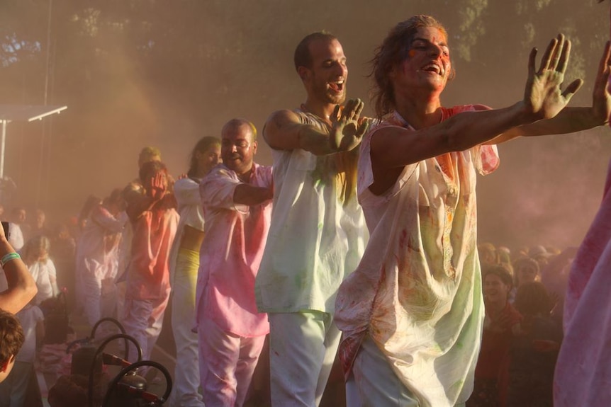 A line of men and women in white clothes, covered in coloured powders.