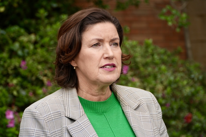 Jackie wears a green top and brown jacket as she stands in an outdoor setting.