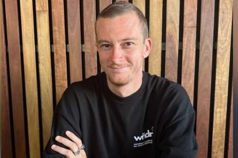 Brett Mckay smiles as he sits arms crossed at a wooden table.