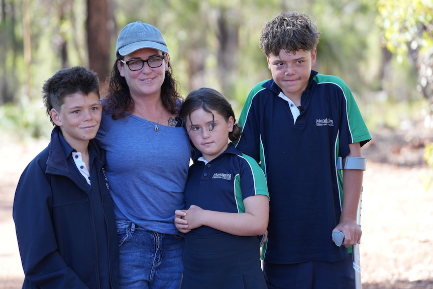 A mother, her two sons and daughter pose for a family photo smiling standing outside in front of bush.