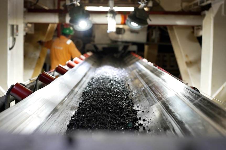 A conveyor belt with fragments of black rocks, stretches away from the camera, in an industrial setting.