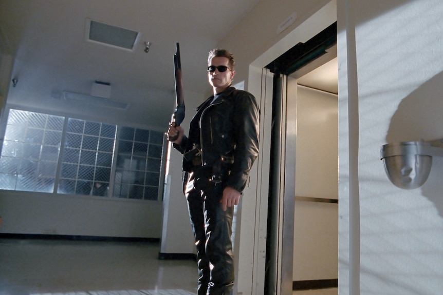 A man in leather pants and jacket holds a big gun in a hallway.