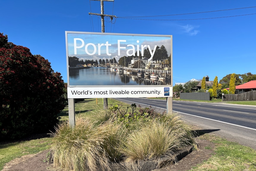 A photo of Port Fairy's town sign on a sunny day, bushes planted underneath, a road beside it, house with red roof in distance.