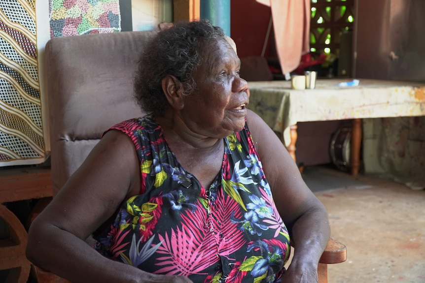 A woman sits on a chair wearing a floral dress and looks slightly off-camera.