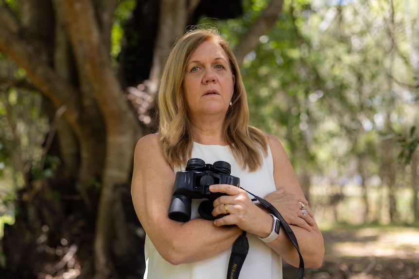 A blonde woman holding binoculars standing under a tree.