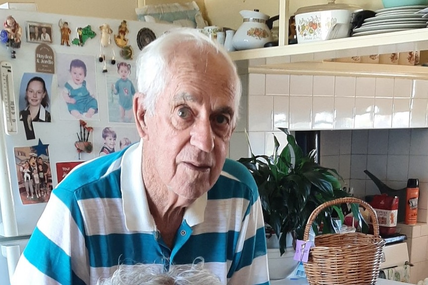An elderly couple, the woman sitting in a chair, the man standing, looking towards the camera inside their home