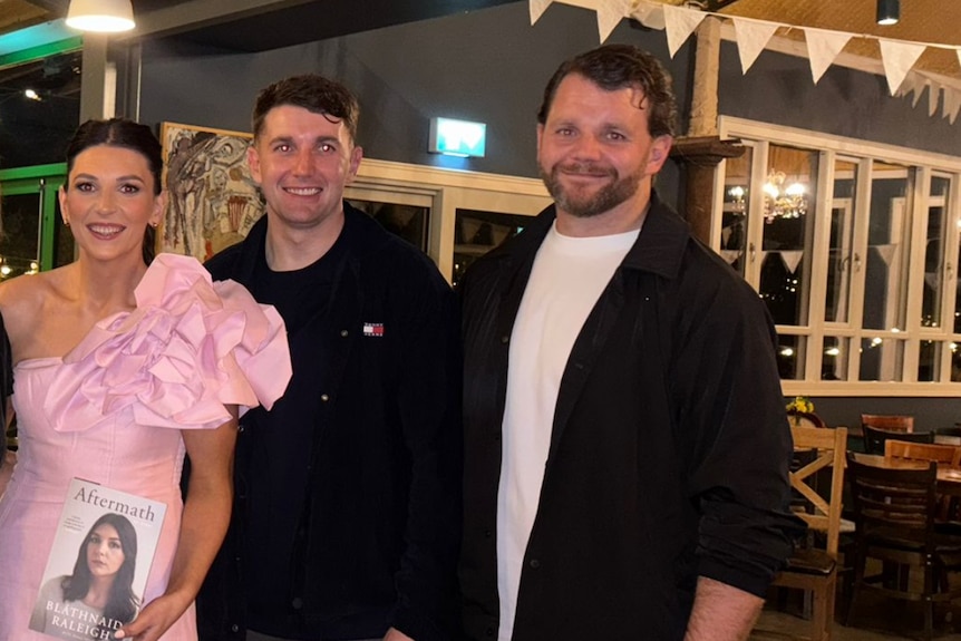 A woman in a pink dress poses with two men while holding a book showing her face on the cover.