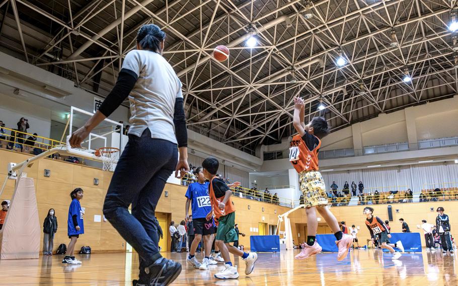 A small boy in an orange jersey takes a jumpshot while teammates and opponents move across the court during a youth basketball game inside a large gymnasium. 