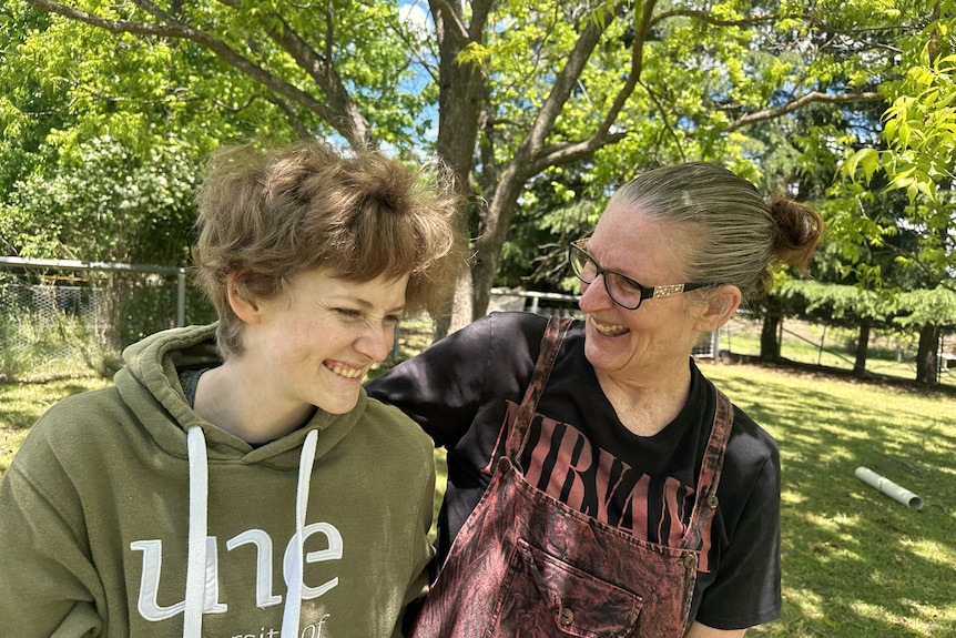 A woman wearing glasses has her arm around a teenager, sitting outside, while both are laughing. 