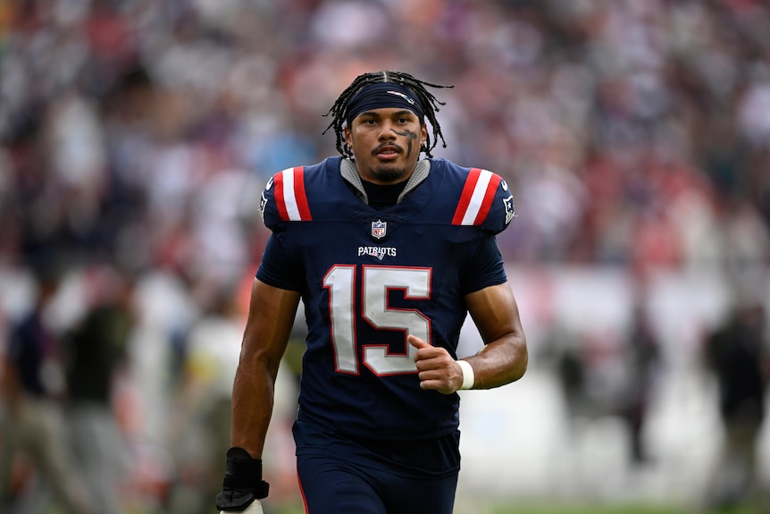 A Samoan man with braids wearing a New England Patriots NFL uniform with the number 15 on the front walks towards the camera 