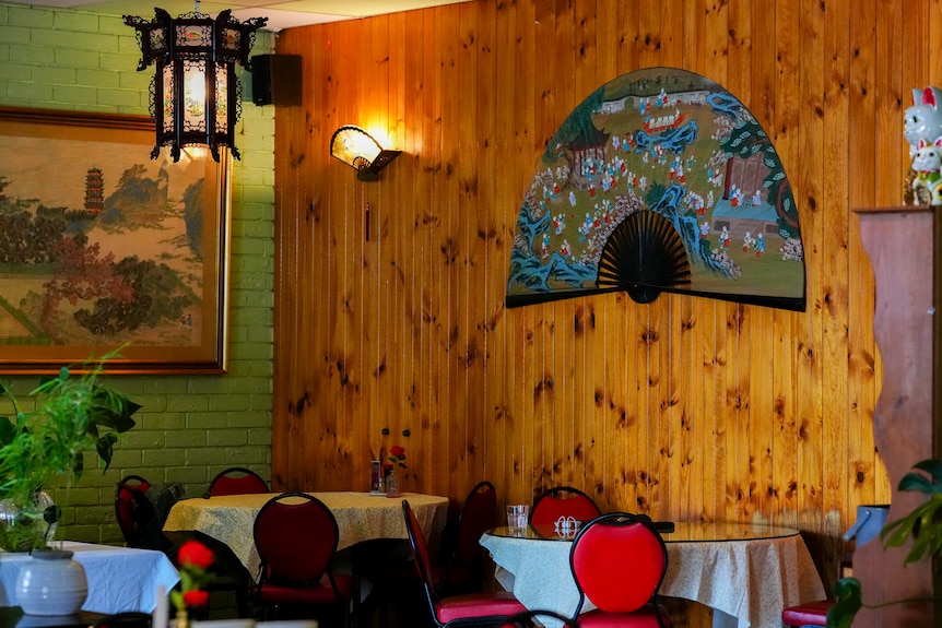 A large oriental fan hung on a wooden wall inside what looks like a restaurant. Empty tables and chairs can also be seen.