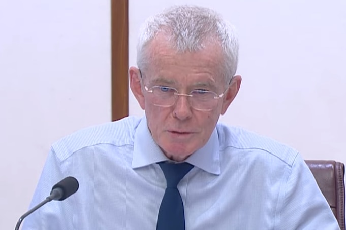 An elderly man with a stern expression talks into a microphone.